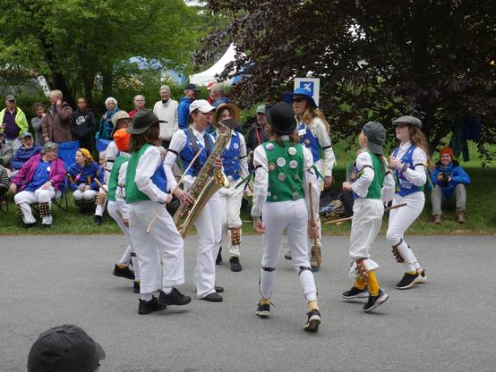 Eight young morris dancers with white shirts and trousers and green and black vests, decorated with badges, dancing on a paved surface. They are surrounding a musician playing a baritone sax. Behind them is an audience including a number of other morris dancers.