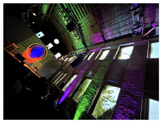 This is the event space at Pioneer Works, a converted factory and arts center in Red Hook, Brooklyn, which recently hosted a panel discussion on UFOs, exoplanets, astrobiology, and the search for extraterrestrial life — a fascinating and wide-ranging conversation between scientists Adam Frank, David Kipping, and Janna Levin (I learned a lot!). I took this photo before the panelists took the stage, when the screen was displaying quirky graphics inspired by the retro “little green men” iconography of the past decades.

Image description: Photo, taken at an askew angle, of a large, darkened hall with multicolored ambient lighting and a glowing graphic projected onto a screen behind a stage. Two rows of windows — taller ground-level ones and smaller upper-story ones — lead from the foreground at right frame to the more distant stage at left frame. Purple and green lighting brings out the texture of the brick columns between the windows, through which can be glimpsed some trees and a pale sky. On the paneled ceiling, which is also lit in green and purple, a row of spotlights illuminates the stage. Onstage are some empty red chairs; behind them, a large screen displays an image of a humanoid silhouette with glowing eyes inside concentric red and yellow circles against a green grid. Above the screen are three small rectangular windows, and above the central window is a circular one. In front of the stage is an audience waiting in shadow.