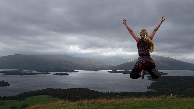 Dieses Foto zeigt vorne rechts der Mitte eine springende Frau von hinten mit ihren Armen weit nach oben ausgestreckt.  Unter ihr ist ein Streifen Grass, dann ein See, dahinter die schottischen Highlands und dadrüber sehr viele dicke, graue Wolken.