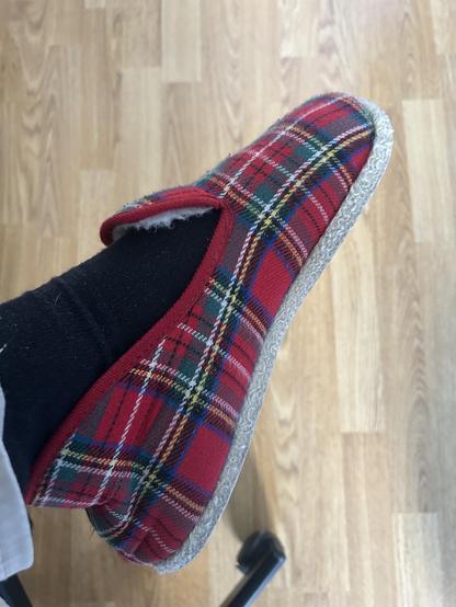 Photo d'une charentaise m'appartenant. Elle a un fond rouge et des croisements de lignes jaunes, blanches, vertes. Je porte en même temps des chaussettes noires. Derrière, on peut apercevoir mon parquet, on ne peut plus classique.