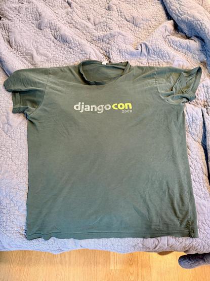 A dark green t-shirt lying on a bed with a blue, textured sheet. The shirt is worn and faded. The neck is starting to fray, and there are small holes visible at the seams. Visible in very faded screen printing on the front are the words ‘djangocon’, with ‘django’ in white lettering, and ‘con’ in a bright green; below that in smaller type in white lettering is ‘2009’.