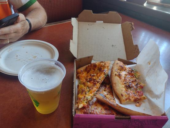 A photo of a open pizza box on a table.  Several slices are stacked in the box and one of them is half-eaten.  Next to the box is a plastic glass of beer with a slim white foam on the top surface.  In the background is an empty almond color paper plate and hands holding a cell phone.