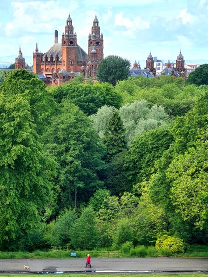 An ornate red sandstone Victorian building rising above the trees of a nearby park.
