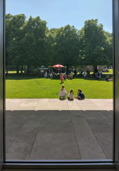 Uitzicht door een venster op het gazon van Museum Voorlinden in Wassenaar, met een ijscowagen, een rood-witte parasol en drie zittende kinderen ¦ View through a window on the lawn of Museum Voorlinden in Wassenaar, with a ice-cream cart, a red-white parasol and three sitting children