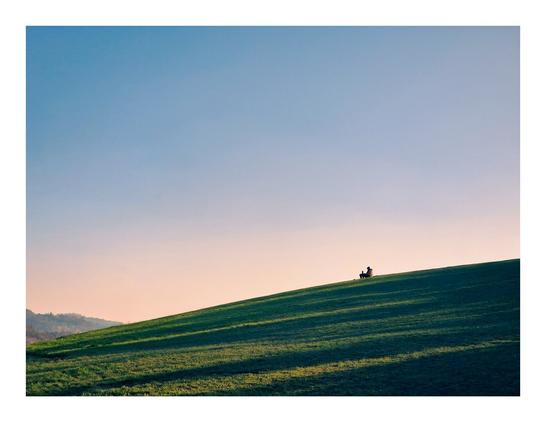 The photo is of a wide green, gently sloping grassy hill. The hill is highest on the right hand side of the photo and slopes down to the left. It is in the evening around sunset. Near the top of the hill, a single person is sitting with a book open on their lap. They are a silhouette against the clear the sky. Long shadows stretch across the hill, from sunlight coming through trees close by but not visible in the photo.