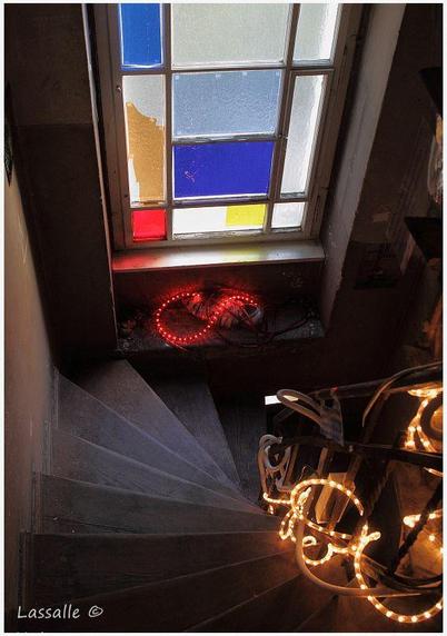 Ein Treppenhaus bzw. Treppenaufgang von oben aufgenommen. Ein typisches Berliner Flurfenster ist mit unterschiedlich farbigem, durchsichtigem Papier beklebt. Unterhalb in der Fensternische leuchtet rot eine Lichterkette. Das rechts der Treppe angebrachte Gelände wurde ebenso mit einer recht langen, weiß gedimmt leuchtenden Lichterkette schlangenförmig versehen. Der Treppenaufgang fällt insgesamt sehr schmal aus.