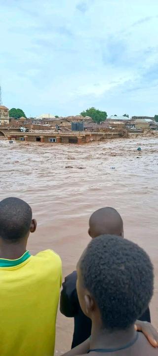 People watching floodwaters across a wide road with water half way up the windows of homes the other side.  image @yaxmokwa Twitter.