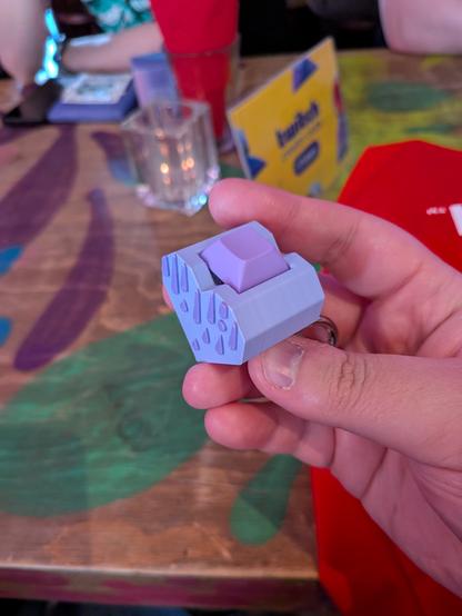 A custom mechanical keycap held in hand, featuring two shades of purple with raised triangular patterns on the sides. The keycap sits atop a heart-shaped base. In the background, a colorful table displays abstract patterns, alongside a glass with a candle, a red napkin, and a card indicating the table is for the Twitch Creator Club for VTubers. The scene has a warm and vibrant atmosphere, filled with thoughtful design elements.