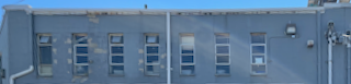 A broad horizontal view of the top floor of an industrial building, gray-painted concrete, the paint chipped and marred, with 7 long, narrow windows across, each window divided vertically into 4 panes. Three of the windows have what could be vent flaps open at the top, others have blinds all the way up or partially drawn, several with gray dish-like objects sitting on the sills, and one has what could be a sheet of paper affixed to the inside. There are drain pipes from the roof at either side, and one between the fourth and fifth windows. It ain't pretty, folks.
