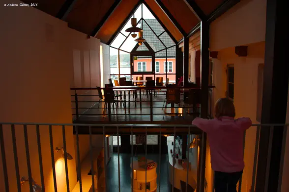 Aufnahme aus dem Inneren der Phänomenta in Flensburg. Ein junges Mädchen steht am Geländer und schaut nach unten. Dort stehen auf einem Tisch Spiele aus dem Angebot des Museumsshops. Auf einer Empore über dem Shop stehen Tische und Stühle eines Cafés, dahinter ein großes Fenster, das über beide Stockwerke geht und den Blick auf das gegenüberliegende Haus freigibt.