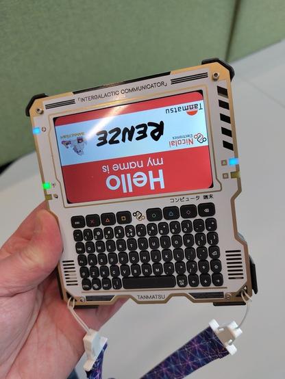 Photo of a handheld computer with a large screen, a qwerty keyboard and dedicated game keys. Wait! No, it's a badge. It even has an MCH strap attached to it. A badge which has _seen things_ and lived to tell the tale.