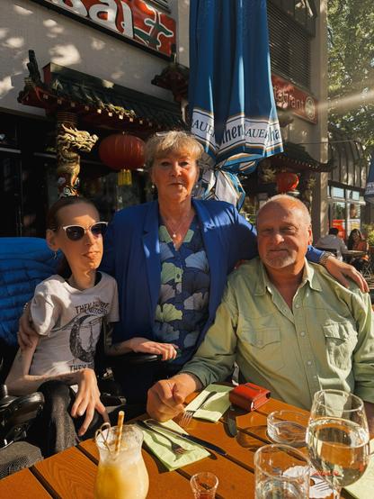 Ich, meine Mutter und mein Vater sitzen und stehen gemeinsam an einem sonnigen Restauranttisch im Freien. Links im Bild sitze ich, mit Sonnenbrille und einem T-Shirt mit dem Aufdruck „They did´nt burn witches, they burned women“. In der Mitte steht meine Mutter mit kurzem blonden Haaren, einem blauem Blazer und geblümter Bluse, die mich und meinen Vater liebevoll berührt. Rechts sitzt mein Vater mit grauem Bart und hellgrünem Hemd. Auf dem Tisch stehen Getränke, Besteck und Gläser. Im Hintergrund sind ein chinesisches Restaurant mit Drachenverzierung und große Sonnenschirme zu sehen. Die Szene wirkt warm, familiär und innig.