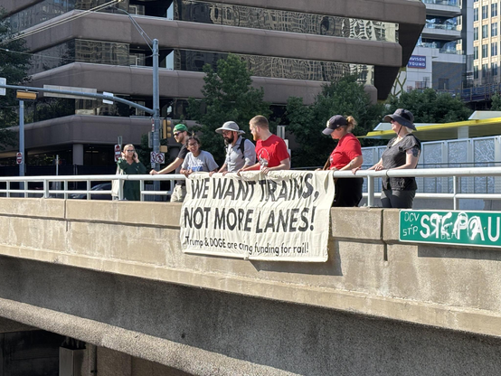 Group of people holding a banner over a highway: 

WE WANT TRAINS, 
NOT MORE LANES!

Trump & DOGE are cutting funding for rail