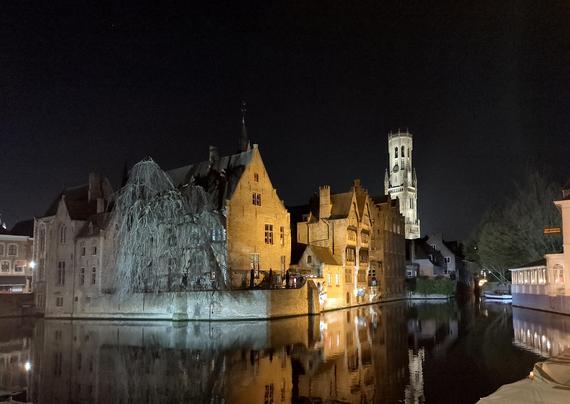 A nighttime view of Bruges, taken from Rozenhoedkaai, over the canal to the towering Belfort, floodlit in white light, contrasting with the golden tones of the buildings of Wollestraat, across the water. Much of the city dates between the 12th and 15th centuries, and the architecture looks ancient and beautiful, with windows and doors almost touching the canal water.