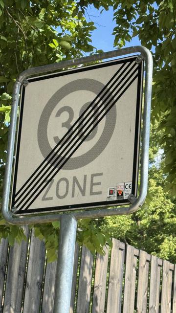 Verkehrsschild (weiß/grau) in Metallrahmen vor Holzzaun und grünbelaubten Zweigen von 2 Bäumen: Zone 30 Ende