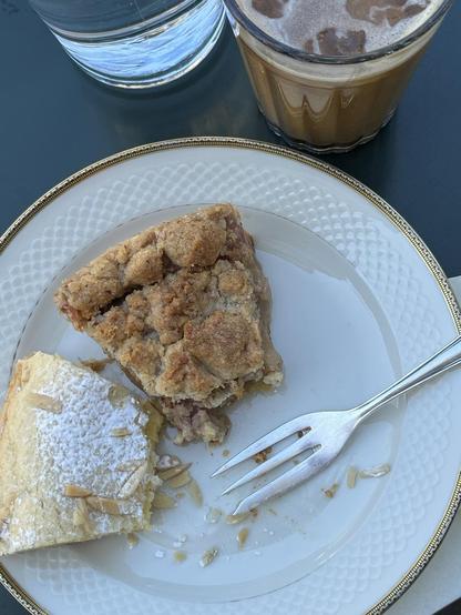 Ein schattiger Café-Tisch mit einem Goldrandteller und zwei angefangenen Kuchenstücken, einem Glas Eiskaffee und einem Glas Wasser.
