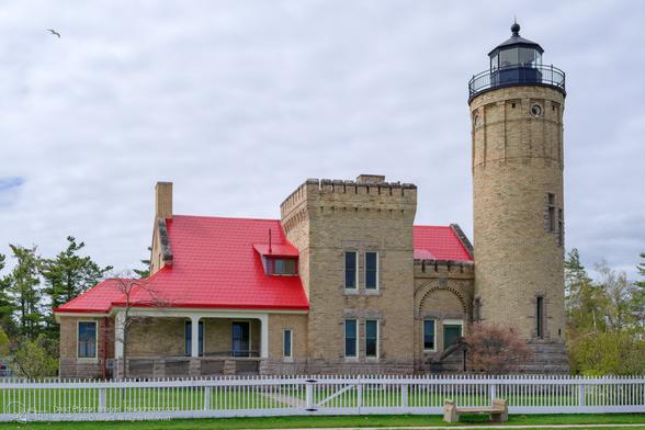 A photograph of the historic Old Mackinac Point Lighthouse, a tan brick building with a bright red roof and a tall cylindrical tower. It stands behind a white picket fence on a cloudy day. A seagull flies overhead.