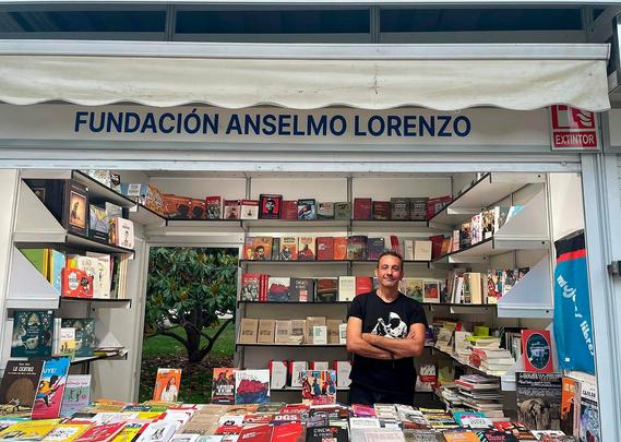 Una persona de pie con los brazos cruzados rodeada de libros en la caseta 55 de la FAL en la feria del libro de Madrid