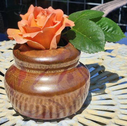 Photo of a brown vase with a gentle design on a white metal table. The vase holds an orange flower with green leaves.