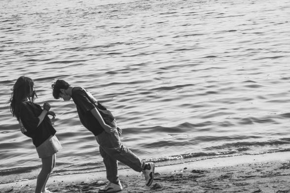 Two people standing on a sandy beach near the water. The person on the left appears to be holding something and the one on the right is leaning towards them. The water reflects light and extends to the horizon. The image is in black and white.