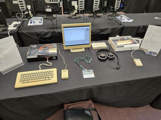 A table covered with a black fabric cover. 

There is a sign including a description of the system to the far left. To the right of the sign is the Macintosh plus clone (a small black acrylic box with a transparent sheet to show the gubbins) with a M0110 and M0100 original Macintosh mouse and keyboard in front. To the right is a teeny VGA monitor from packard bell, showing the system has booted up. 

In the middle is my DosFox name tag. 
To the right is a small beige box, with a set of headphones attached - this is a Yamaha MU10 midi synth with a pair of AKG headphones.

To the right of those is the Unitron 1024, with a single Macintosh mouse plugged into it - this is an identical box to the Mac plus, except it's in white and says "Unitron 1024".

To the extreme right of that is a floppy emu, and another information sign for the Unitron 1024.