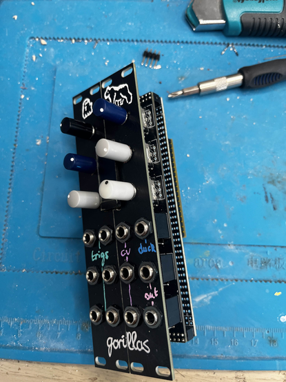a prototype Eurorack module. there are six rotary encoders around a small OLED screen, and twelve jack sockets, six labelled "trigs", three labelled "CV", two labeled "out" and one labelled "duck". the module is constructed from stacked boards a little more sophisticated (but not much) than protoboard. the front panel labels are in multicolour pastel paint marker. the module name is "Gorillas" in cursive handwriting, and there's a drawing of a gorilla and a pale slim ghost as logos.