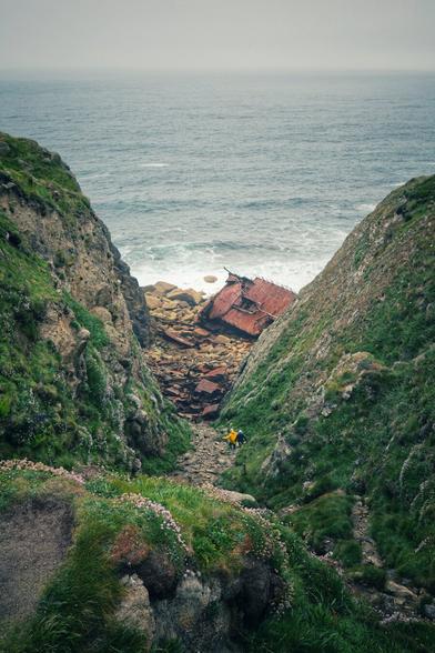 Looking down a gully between two steep rock walls, towards a beach and the sea. On the narrow section of beach visible at the end is a rusted hull of a ship. Two small figures can be made out walking along the gully.