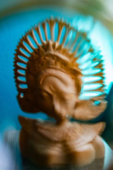 A distorted photo of a wooden Tara goddess bust. It appears that her head is bending forward slightly and her facial features are askew. There is an aqua blue wall in the background and some highlights in the upper right corner from a window. End description.