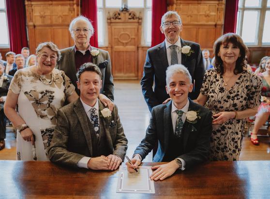 Photo of me and my now-husband sitting at a big wooden desk with our parents standing behind us. We're all looking at the camera and smiling. I'm holding a pen in my hand over a ceremonial wedding certificate.