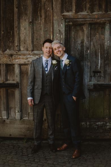 Wedding photo of me and Ady in our suits - me in a dark blue, him in grey - standing in front of a huge wooden door in Oxford. We're both smiling at the camera and look very happy.
