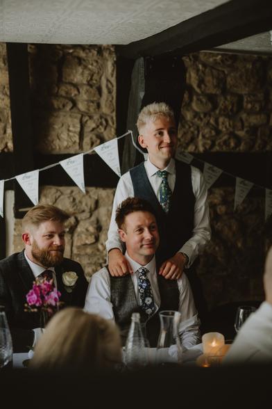 Candid photo of us taken at the wedding reception where we're both listening to a speech that's being given off-camera to the right. Ady is sitting at the table while I'm standing behind him with my hands on his shoulders.