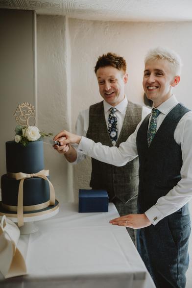 Photo of me and my husband cutting our wedding cake together. The cake is fairly simple - blue, two tiers, with a gold ribbon around it and a small white rose on top. We're wearing our suits still, but have taken the jackets off to show our waistcoats instead.