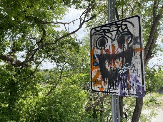 In the background blue skies, muddy river and green trees. In the foreground more green trees. In front of all that closest to us, at the right of the frame, an abused tin sign on a pole. It has been painted and painted over numerous times with people’s names and phrases in different colours. There’s the vague remnants of a bicycle from the original sign that have been turned into eyes; a crazed wide open black painted dripping mouth below creates a manic visage.
