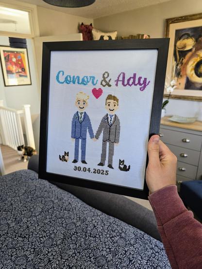 Photo of me holding out a cross-stitch done by my friend Hayley. It shows me and my husband in cute, almost pixel art style, holding hands wearing the suits we wore at our wedding, right down to the tie colours. Next to us in the cross-stitch are representations of our two cats, one either side of us, again looking very cute. And at the bottom is the date of our wedding, 30 April. In the background you can see one of the cats and it's clear how good a likeness it is!