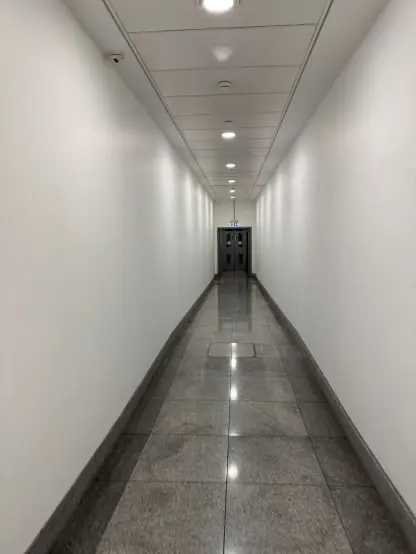 A long, plain corridor, with white walls, stone floors, and panelled ceiling, with clean cold white lighting. The corridor is slightly descending.