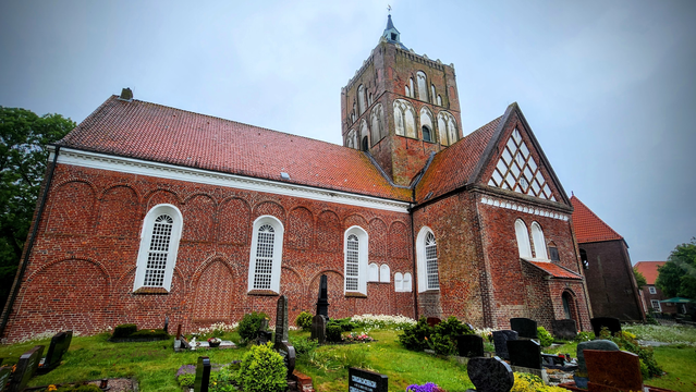 Eine ziegelsteinrote Kirche mit Querschiff hat einen mächtigen Turm, der aufragt. Vor der Kirche sind Gräber zu sehen.