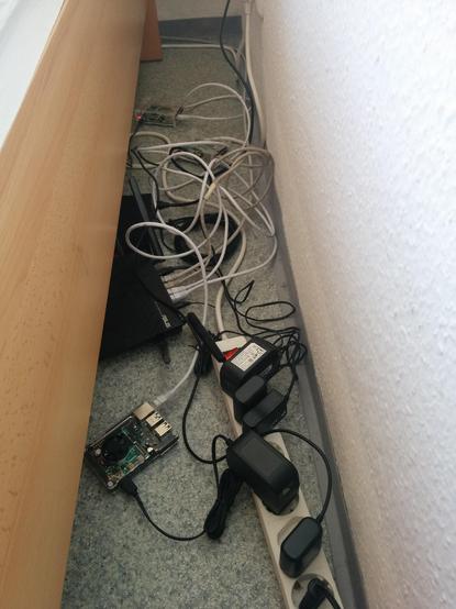 Ein Foto von dem Spalt zwischen einem Bett und einer Wand. Das Bett wurde scheinbar etwas vorgezogen sodass zu Vorschein kommt, was unter dem Bett liegt.

Es ist eine lange Steckerleiste mit vielen Netzteilen und Kabeln. Dazwischen ein WLAN Router, Switch und mehrere Raspberry PIs.