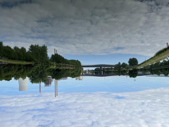 Cool power plant, a Bridge and some more industry buildings, also some trees, all reflected in a canal. Above it a Bit of Blue sky shining through a lot of white clouds. The Picture is turned upsidedown.