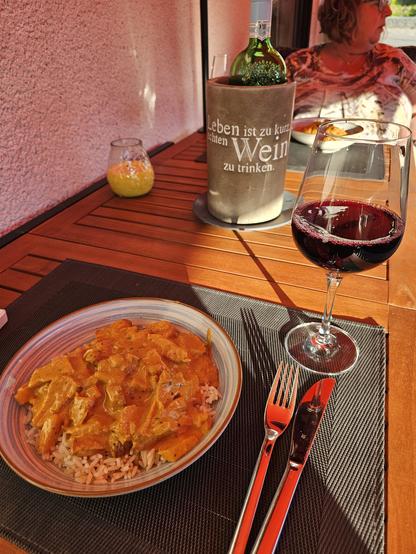 Das Bild zeigt eine gemütliche Mahlzeit an einem Holztisch. Im Vordergrund befindet sich ein Teller mit einem Gericht, das aus Reis und einer cremigen, orangefarbenen Soße besteht, die wahrscheinlich eine Art Curry ist. Neben dem Teller steht ein Glas Rotwein, das halb gefüllt ist. Ein Besteck, bestehend aus einem Messer und einem Gabel, liegt daneben. Im Hintergrund ist eine Flasche Wein in einem Eisbehälter zu sehen, auf dem der Text "Das Leben ist zu kurz, um schlechten Wein zu trinken" steht. Ein weiteres Glas mit einer Kerze gegen Mücken steht ebenfalls auf dem Tisch. Im Hintergrund sitzt eine Person am anderen Ende vom Tisch. Die Szene wirkt entspannt und einladend, mit warmem Sonnenlicht, das den Tisch beleuchtet.

Bereitgestellt von @altbot, privat und lokal generiert mit Ovis2-8B
