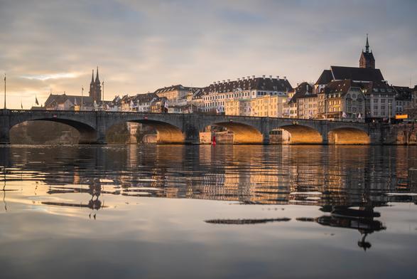 Am Rhein in Basel, Blick auf die historische Mittlere Brücke und Grossbasel. Der Himmel ist größtenteils bedeckt, aber links außerhalb des Bildes scheint die Morgensonne durch eine Wolkenlücke und beleuchtet die Brückenbögen von unten. Auch die Gebäude im Hintergrund liegen im warmen Sonnenlicht. Das Ganze spiegelt sich im Rhein, wobei die Spiegelung durch Wellen verzerrt ist.