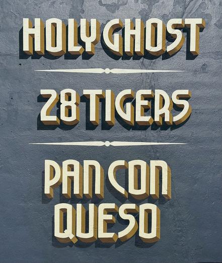 A hand-lettered sign painted on the side of a building: light yellow letters with deep gold edges appear to cast realistic shadows on a dusty blue background. The sign lists three businesses: “Holy Ghost, 28 Tigers, Pan Con Queso”