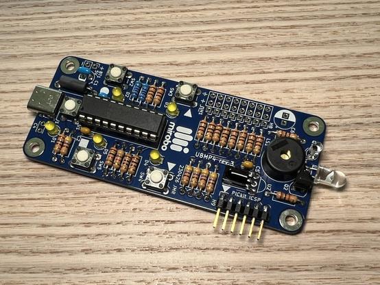 A fully assembled UBMP4 circuit sitting on a wooden tabletop. From left to right, the circuit features a USB type-C connector, a PIC16F1459 microcontroller surrounded by 5 pushbuttons and 5 LEDs, an expansion header area, an IR demodulator, an in-circuit programming header, a piezo speaker, an ambient light sensor, and an IR LED.