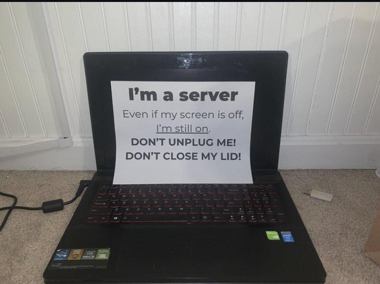 An old laptop that has a white label on the screen that says: "I'm a server. Even if my screen is off, I'm still on (underlined) DON'T UNPLUG ME! DON'T CLOSE MY LID!"
