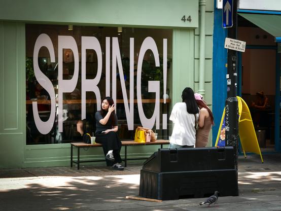 Three young women, two seen from behind standing next to a sand/salt bunker near the kerb and one seen from in front sitting on a long wooden bench outside a shop window in which is a large poster with the word, "SPRING!". It's the second to-last day of the last Spring month, but this was taken earlier.