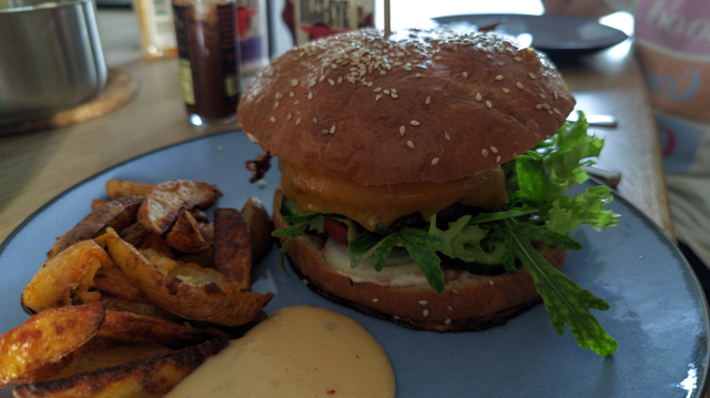 ein großer Burger aus dem etwas Rucola herausragt, Kartoffelecken und Chilicheesesauce auf einem blaugrauen Teller