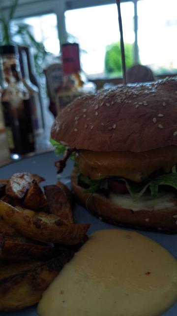 ein großer Burger aus dem etwas Rucola herausragt, Kartoffelecken und Chilicheesesauce auf einem blaugrauen Teller