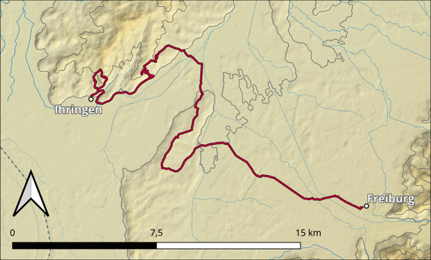 Das Bild zeigt eine topografische Karte, die eine Route von Ihringen nach Freiburg darstellt. Die Route ist in Rot markiert und verläuft durch eine hügelige Landschaft. Die Karte zeigt die Höhenlinien, die die topographische Struktur der Region veranschaulichen. Die Route beginnt in Ihringen im Nordwesten und führt nach Süden bis nach Freiburg im Südosten. Ein Kompass in der linken unteren Ecke zeigt die Richtung Nord. Eine Skala am unteren Rand der Karte zeigt die Entfernung in Kilometern, wobei 7,5 km und 15 km markiert sind. Die Karte ist in einem hellen Beige-Ton gehalten, mit blauen Linien, die Flüsse oder Gewässer darstellen.