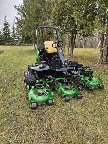 A large ride-on lawnmower that consists of a four-wheeled vehicle with driver seat, steering and other controls. Attached to the front of it are three green lawn mowers with their own wheels. Where the engine would sit, a hydraulic motor is mounted on each, with hoses going to the main vehicle. It looks like the big machine is pushing three regular manual lawn mowers in parallel.