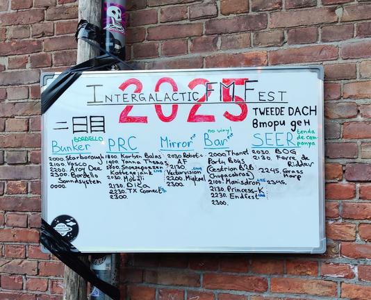 Partial DJ/live timetable scribbled on a whiteboard duct taped to a pipe on a brick wall