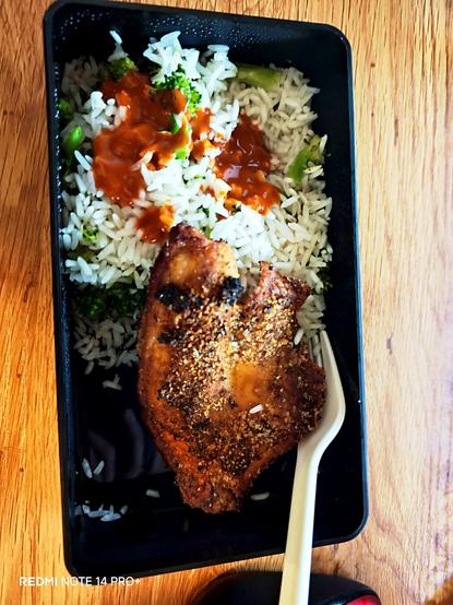 A bento box of rice and broccoli with a few drops of hot sauce. A fried tilapia. A fork sticking out of the box and the box is placed on a wooden desk.